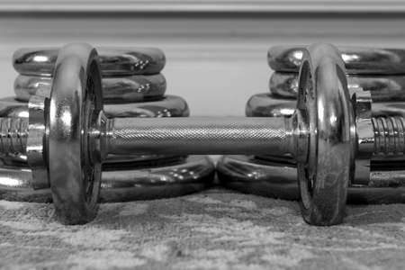 Black and white photo of a small dumbbell on the floor with two stacks of weights behind itの写真素材