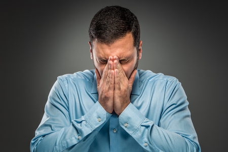 Closeup portrait of upset man in blue shirt and butterfly tie covering his face by hands,  isolated on gray background. Feeling sick and tired - Human emotion.の写真素材
