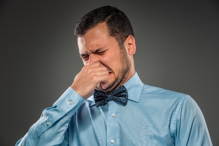 Young man in blue shirt and butterfly tie, with disgusted expression closing his nostrils with his fingers, isolated on gray background. Closeup. Mimicry. Gesture.の写真素材