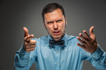 Portrait of angry upset young man in blue shirt and butterfly tie with hands up, pointing finger at camera, isolated on gray background. Negative human emotion, facial expression. Closeupの写真素材