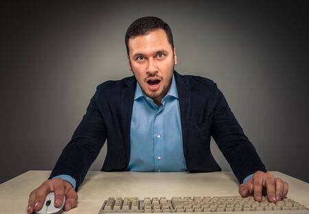 Portrait young man in blue shirt and jacket looking at the camera with amazement, sitting at a desk...