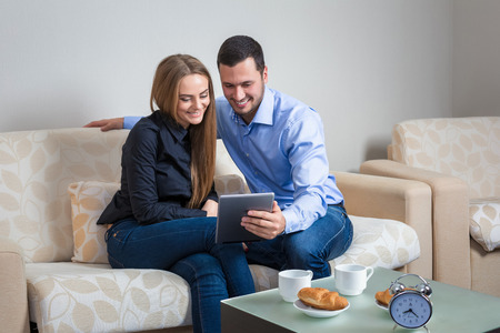 Beautiful young couple laughing,  bvsitting on a sofa, sharing photos or other information, displayed on an electronic tablet with each other and drinking coffee with croissantsの写真素材