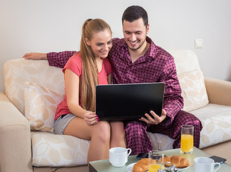 Cute young couple on the couch having breakfast, eating croissants, drinking orange juice  in front of a laptop computerの写真素材