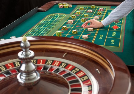 Close up view of Roulette and piles of gambling chips on a green table in casino. Man hand over...