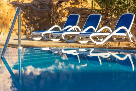 Three sunloungers near beautiful swimming pool and reflected their in a blue water of the pool....