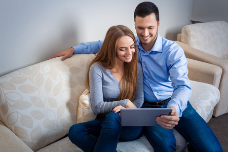 Beautiful young couple laughing, sitting on sofa, sharing photos or other information, displayed on an electronic tablet with each otherの写真素材