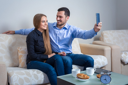 Young man and woman doing selfie with telephone camera, happy people taking picture smiling to the camera, sitting on sofaの写真素材