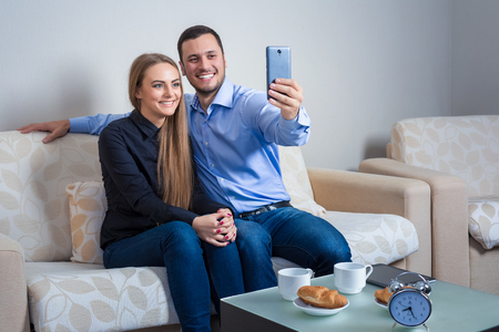 Young man and woman doing selfie with telephone camera, happy people taking picture smiling to the camera, sitting on sofaの写真素材