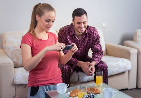 Cute young couple on the couch having breakfast, eating croissants, drinking orange juice. Woman photographs breakfast table at mobile phoneの写真素材