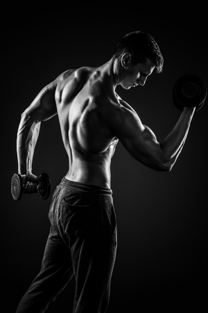 Fitness man working out doing exercises with dumbbells at biceps on black background, rear view. Black and whiteの写真素材
