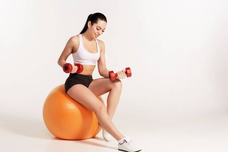 Young beautiful sportswoman in white top and black shorts with dumbbells exercising on fitball.の写真素材