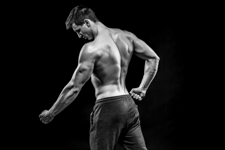 Image of very muscular man posing with naked torso in studio and looking behind on black background. Black and whiteの写真素材