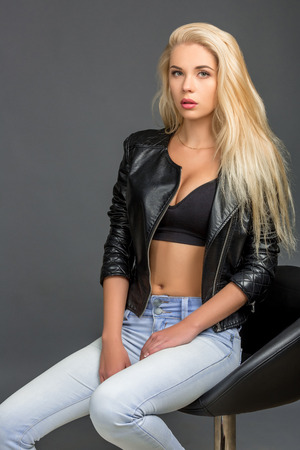 young blond woman in a black leather jacket, blue jeans and a top. she is sitting on a chair and looking at the cameraの写真素材