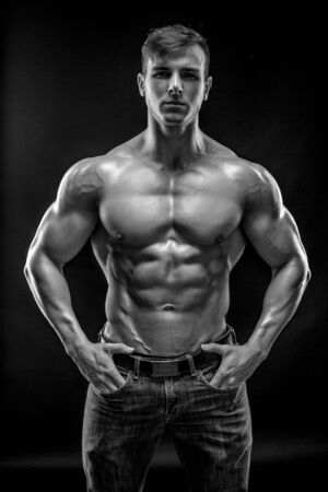 Muscular bodybuilder guy doing posing over black background. Naked torso in jeans. He looks into the camera. Black and white, b wの写真素材