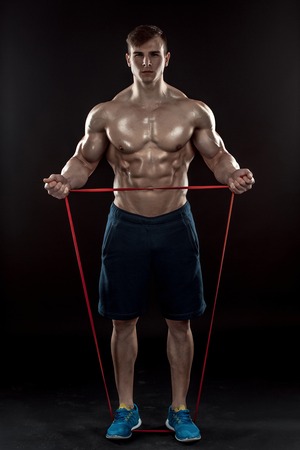 Young athletic man exercising and doing fitness with a chest expander, resistance band, on dark...
