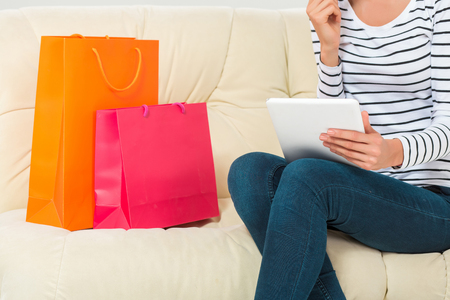 Online shopping and technology concept - smiling beautiful young woman with tablet pc computer and credit card. She sit on sofa with colorful bagsの写真素材
