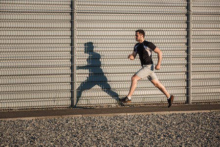 Full length portrait of athletic man running along beautiful grey wall outdoors, muscular build young runner working out while jogging in the park. Sunsetの写真素材