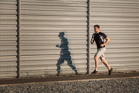 Full length portrait of athletic man running along beautiful grey wall outdoors, muscular build young runner working out while jogging in the park. Sunsetの写真素材