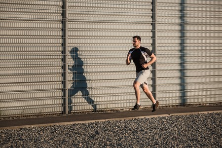 Full length portrait of athletic man running along beautiful grey wall outdoors, muscular build young runner working out while jogging in the park. Sunsetの写真素材