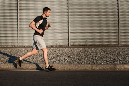 Full length portrait of athletic man running along beautiful grey wall outdoors, muscular build young runner working out while jogging in the park. Sunsetの写真素材