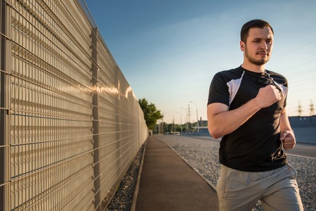 Close-up portrait of athletic man running along beautiful grey wall outdoors, muscular build young runner working out while jogging in the park. Sunsetの写真素材