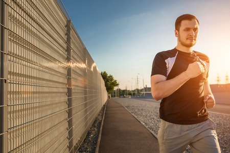 Close-up portrait of athletic man running along beautiful grey wall outdoors, muscular build young runner working out while jogging in the park. Sunset, with solar flareの写真素材