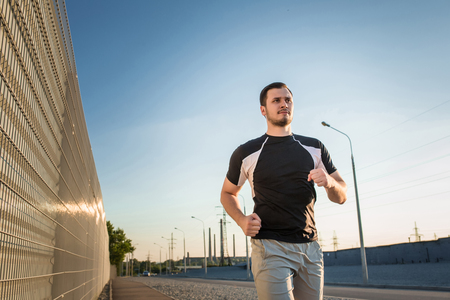 Close-up portrait of athletic man running along beautiful grey wall outdoors, muscular build young runner working out while jogging in the park. Sunsetの写真素材