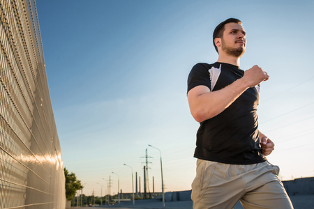 Close-up portrait of athletic man running along beautiful grey wall outdoors, muscular build young runner working out while jogging in the park. Sunsetの写真素材