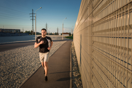 Close-up portrait of athletic man running along beautiful grey wall outdoors, muscular build young runner working out while jogging in the park. Sunsetの写真素材