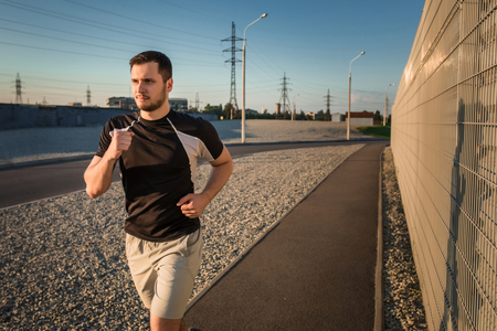 Close-up portrait of athletic man running along beautiful grey wall outdoors, muscular build young runner working out while jogging in the park. Sunsetの写真素材