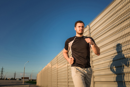 Close-up portrait of athletic man running along beautiful grey wall outdoors, muscular build young runner working out while jogging in the park. Sunsetの写真素材