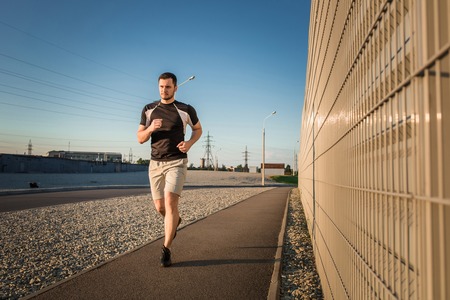 Close-up portrait of athletic man running along beautiful grey wall outdoors, muscular build young runner working out while jogging in the park. Sunsetの写真素材
