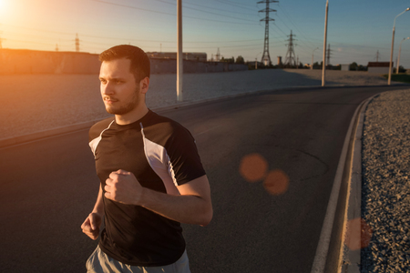 Close-up portrait of athletic man running along beautiful grey wall outdoors, muscular build young runner working out while jogging in the park. Sunset, with solar flareの写真素材