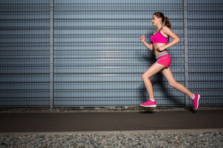 Running woman. Runner jogging against the background of a gray wall. Female fitness model training outsideの写真素材