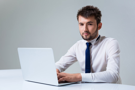 young man smiling at the camera uses a laptop computer while sitting at a table. Office clothingの写真素材
