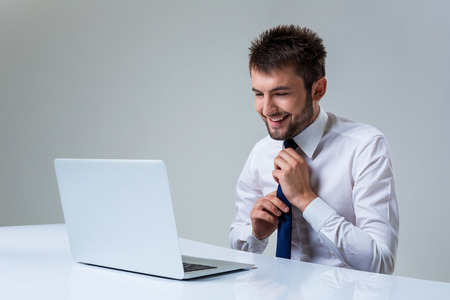 young man smiles and straightens his tie using laptop computer sitting at the table. Office clothingの写真素材