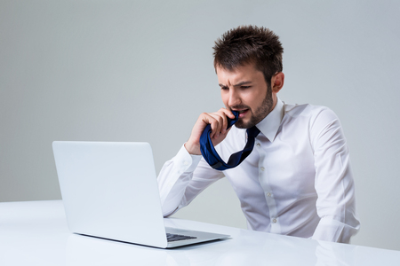 the young man is nervous and chewing on his tie using laptop computer sitting at the table. Office clothingの写真素材