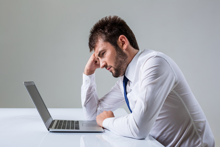 tired young man looking at laptop. It uses a computer while sitting at a table. Office clothingの写真素材