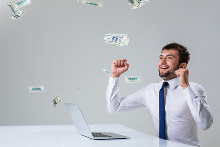the young man behind the laptop. It uses a computer while sitting at a table. enjoys hands up. Office clothing. the top falling money, dollarsの写真素材