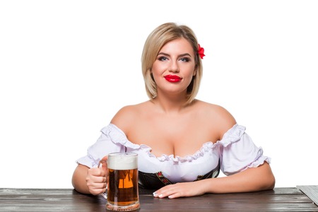 Beautiful young blond girl in dirndl drinks out of oktoberfest beer stein. Isolated on white background. sits by the tableの写真素材