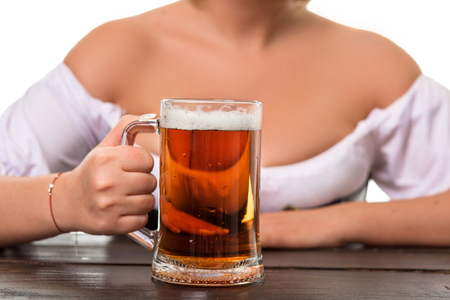Beautiful young blond girl in dirndl drinks out of oktoberfest beer stein. Isolated on white background. sits by the table. close-up mugの写真素材