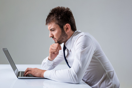 the young man stares at a laptop. It uses a computer while sitting at a table. Office clothingの写真素材