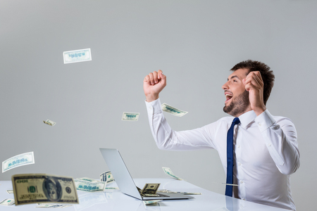 the young man behind the laptop. It uses a computer while sitting at a table. enjoys hands up. Office clothing. the top falling money, dollarsの写真素材