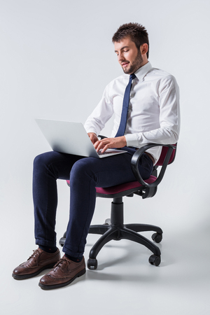 emotional young guy in office clothes working on a laptop computer and sitting on a chairの写真素材