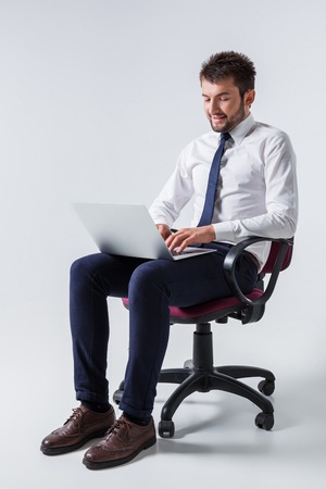 emotional young guy in office clothes working on a laptop computer and sitting on a chairの写真素材