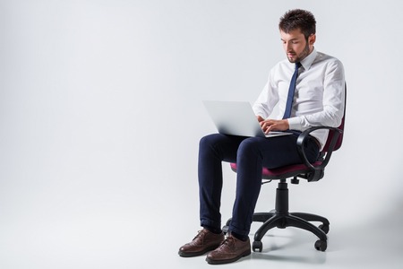 emotional young guy in office clothes working on a laptop computer and sitting on a chairの写真素材