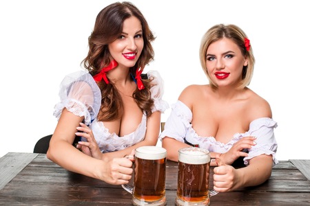 Two beautiful young blond and brunette girls in dirndl drinks out of oktoberfest beer stein. Isolated on white background. sits by the tableの素材 [FY31062816694]