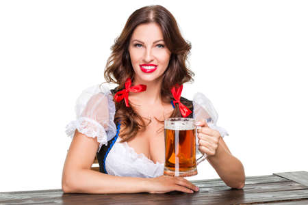 Beautiful young brunette girl in dirndl drinks out of oktoberfest beer stein. Isolated on white background. sits by the tableの写真素材