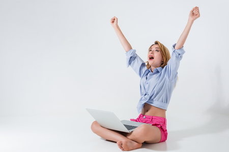 young woman sitting on the floor with crossed legs and using laptop on white background. very satisfied, happy, she raised her hands upの写真素材