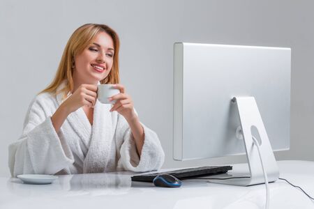 young woman sitting in the table and using computer on gray background. girl smiling and looking at monitor. with a cup in handsの写真素材